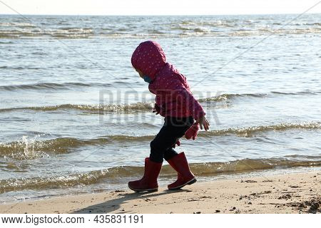 Child Girl Playing By The Sea In Autumn. A Girl In A Red Jacket In A Beautiful Autumn On The Seashor