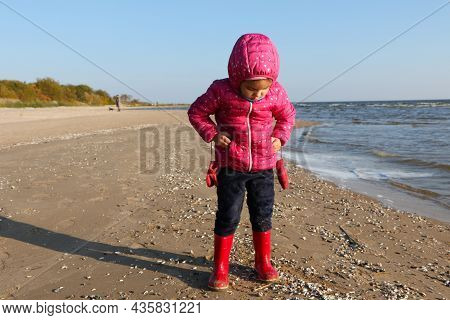 Child Girl Playing By The Sea In Autumn. A Girl In A Red Jacket In A Beautiful Autumn On The Seashor