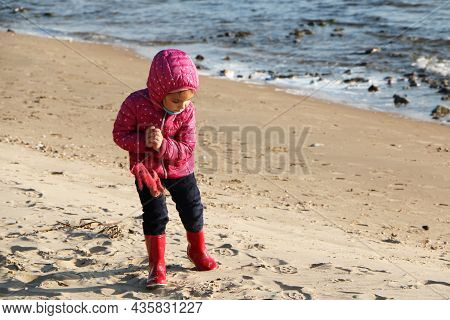 Child Girl Playing By The Sea In Autumn. A Girl In A Red Jacket In A Beautiful Autumn On The Seashor