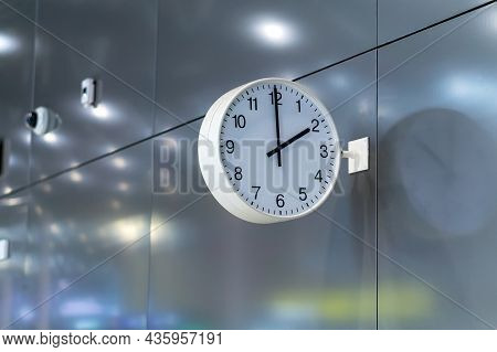 A White Public Clock On A Metal Wall. They Show Two Hours.