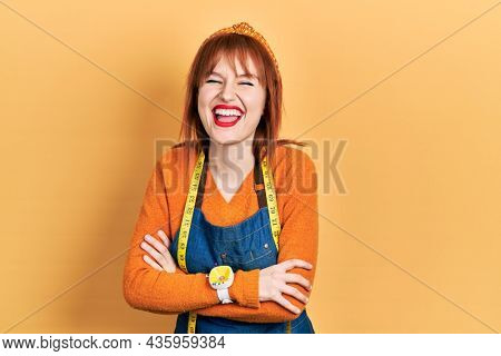 Redhead young woman dressmaker designer wearing atelier apron smiling and laughing hard out loud because funny crazy joke. 