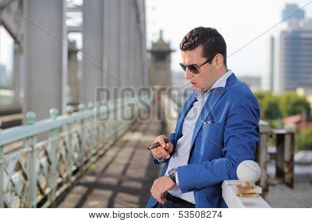 Handsome man in jacket and sunglasses with cigar stands near railings and thinks.