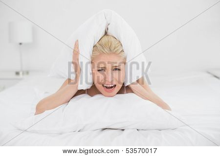 Portrait of an angry young woman covering ears with pillow in bed at home