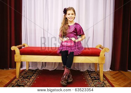 Beautiful girl in dress sits on soft bench near curtains in theatre