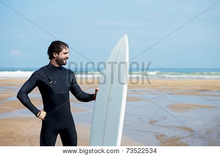 Young professional surfer smiling standing on beautiful ocean beach with his white surfboard
