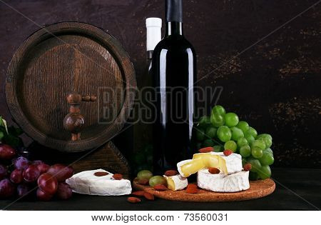 Supper consisting of Camembert cheese, wine and grapes on cutting board and wine barrel on wooden table on brown background