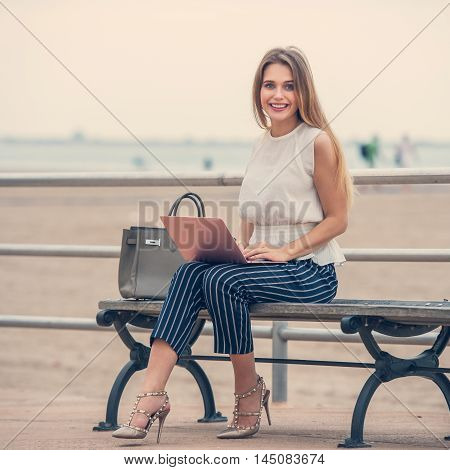Young happy smiling student girl sitting on a bench with a laptop. Cherrful woman working and typing on laptop outdoors.