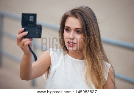 Beautiful girl sitting outdoors and taking selfie video for fashion blog. Woman holding compact camera in her hand ant taking photo.