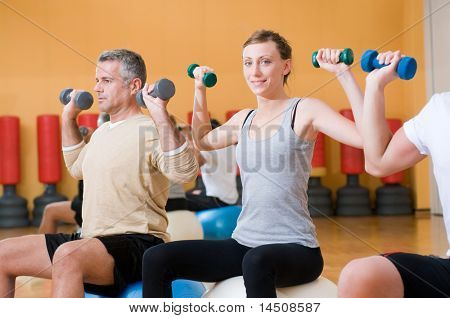 Young lady smiling and looking at camera while lifting weigths on a fitness ball at gym