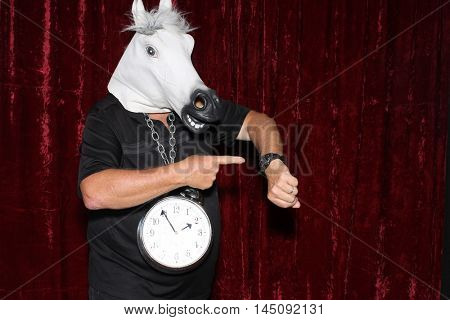 An unidentifiable man wears a rubber Horse Head a Clock around his neck in a Photo Booth.
