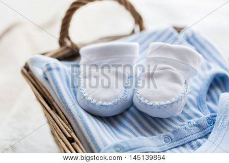 clothing, babyhood, motherhood and object concept - close up of white baby bootees with pile of clothes and towel for newborn boy in basket on table