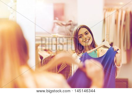 shopping, fashion, style and people concept - happy woman choosing clothes and looking to mirror in mall or clothing store
