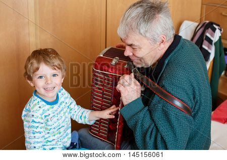 Happy blond little kid boy and his grandfather playing together with accordion. Senior man teaching his grandson, cute toddler to play with music instrument at home.