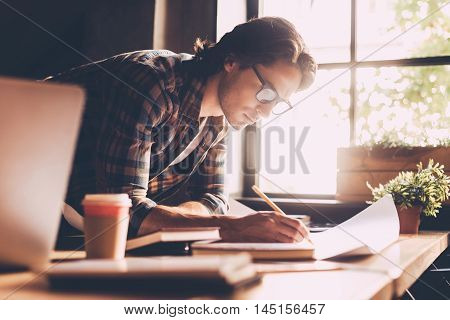 Time to work. Confident young man in casual wear sketching on blueprint while standing near wooden desk in creative office