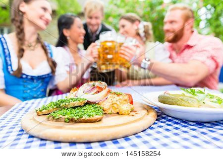 Five friends, women and men, sitting in beer garden clinking glasses having Bavarian appetizers on the table
