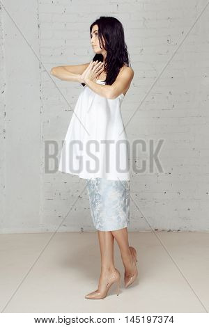 Model in studio with long black hair posing to the photograther like she is praying and looking aside. Studio has grey brick walls. She has nice light costume