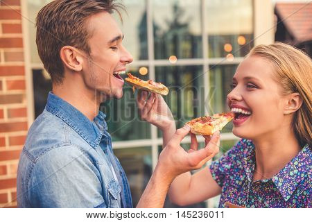 Side view of beautiful young couple eating pizza looking at each other and smiling while walking outdoors