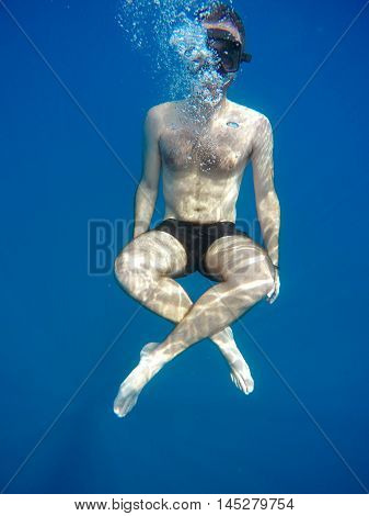 Portrait of a young causasian man meditating in the lotus position underwater. Submerged under water free diving crossed legs wearing a mask and blowing bubbles.