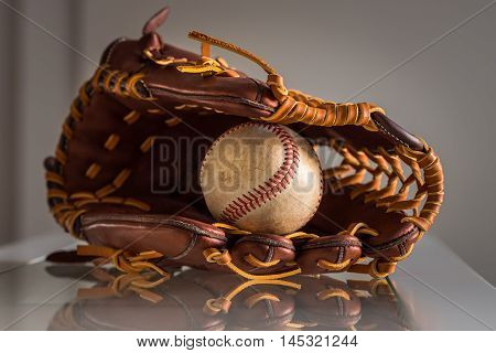 Close-up of a used baseball ball inside brown leather baseball glove on plain grey background.