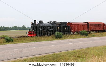 Swedish steam freight train between a road and a field on a cloudy overcast day