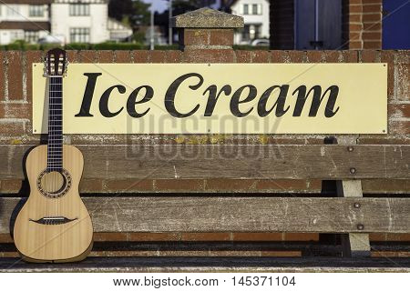 Parlour-sized classical acousic guitar resting on a bench infront of an ice cream sign. Symbolic of vacation relaxation and seaside fun.