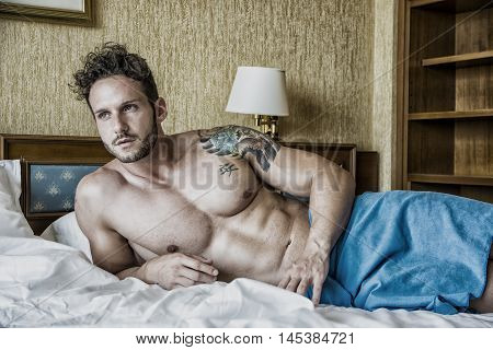 Shirtless sexy male model lying alone on his bed in his bedroom, looking away with a seductive attitude