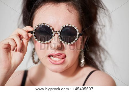 Dark-haired girl model in stylish round dark glasses shows tongue posing in studio.