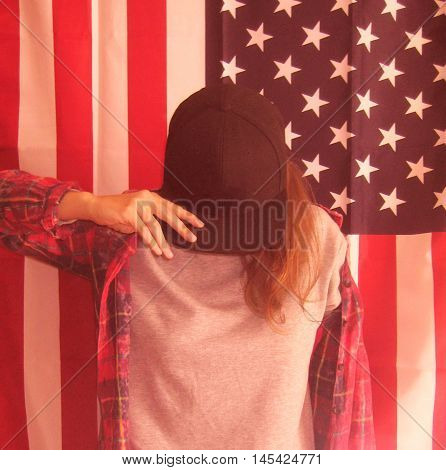 Vintage lifestyle portrait of young hipster girl against american flag background wearing casual clothes gray pullover, red plaid shirt and black snapback cap
