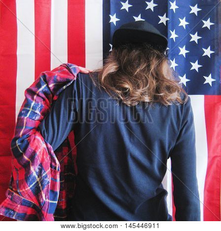 Lifestyle cropped portrait of young hipster girl dressing putting on plaid red shirt getting ready to go out. Casual clothes black pullover and black snapback cap with hair down on american flag background