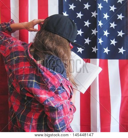 Retro lifestyle portrait of teenage girl reading a book against american flag background wearing casual clothes black plain t-shirt, red plaid shirt, black snapback cap and glasses