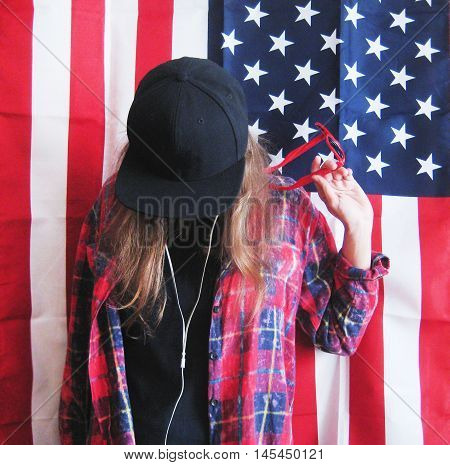 Vintage lifestyle portrait of happy teenage girl standing against american flag background wearing casual clothes black plain t-shirt, red plaid shirt, black snapback cap and holding sun glasses and listening to music