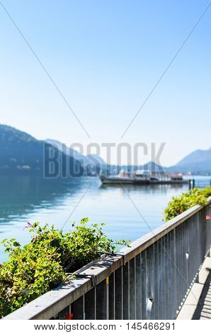 Vacation summer sunny lake view. Beautiful flowers over the background of beautiful serene blue Lugano lake clear blue sky and a ship surrounded by hills in Morcote Switzerland selective focus