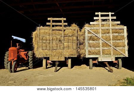 An old orange tractor and two loaded hayracks full of grain bundles are parked in the entrance of a steel shed