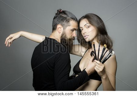 young couple of sexy woman with pretty face and long brunette hair in black bra on body and handsome bearded man visagiste with fashionable makeup brush in studio on grey background