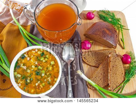 Traditional russian summer cold kvass soup okroshka in a white ceramic bowl jug with kvass (kvas) radish green onion and black rye bread on wooden cutting board. Healthy food concept. Top view. Horizontal.