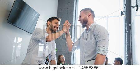 Two businessmen showing victory, unity while communicating in officeinterior. Happy man looking at each other and smiling. Business or freelance concept.