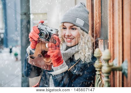 Young blond curly female in warm clothers with old film camera shooting a photo on the background of wooden doors with handles, winter, snow