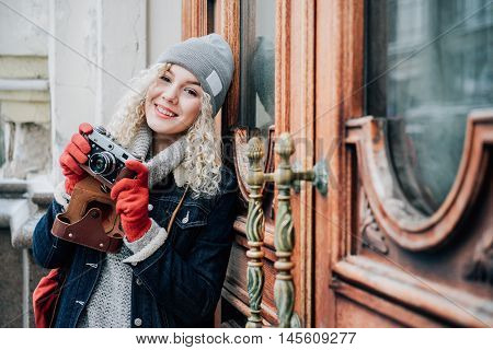 Young blond curly female in warm clothers with old film camera shooting a photo on the background of wooden doors with handles, winter, snow
