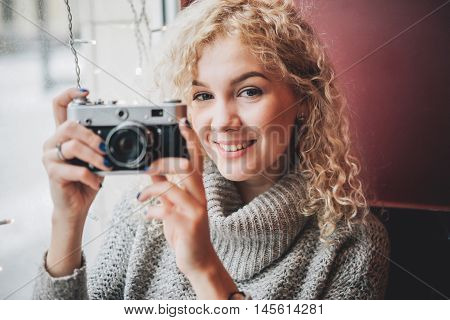 Young blond curly female in warm sweater with old film camera smiling and shooting a photo in the cafe, winter city outside the window