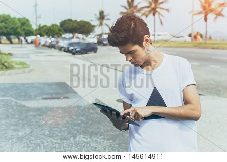 Attractive modern young white male with black hair in white t-shirt using tablet computer, standing on sea promenade with palm trees, sunny bright summer day, vintage color filter