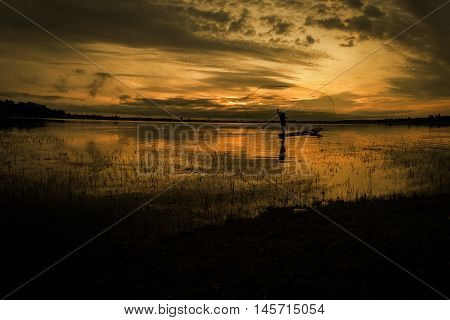 Asian fisherman on wooden boat casting a net for catching freshwater fish in nature river in the early morning before sunrise