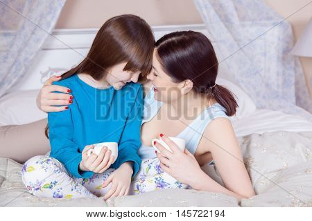 Mother and daughter lay in bed and drinking tea smiling and looking at camera. Studio shot.
