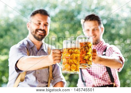 Two handsome hipster young men in traditional bavarian clothes holding mugs of beer, clinking. Oktoberfest. Sunny summer garden.