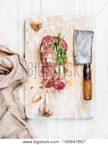 Raw uncooked roastbeef meat cut with rosemary, thyme and garlic and butcher knife on old white painted wooden background, top view, vertical composition