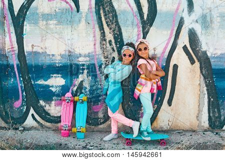 Two 7 years old children wearing cool fashion clothing posing with colorful skateboard against graffiti wall, urban style