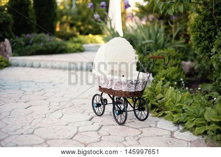 Doll's pram. Vintage doll stroller placed on the stone walkway alley in a beautiful garden with flowers and trees around. Retro cart dolls made of rattan and white lace