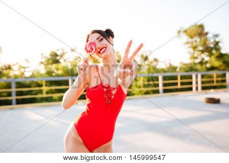 Happy cute young woman covered her eye with lollipop and showing peace sign standing outdoors