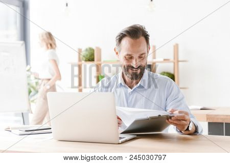 Friendly man manager working on laptop computer with documents while sitting at the office with woman co worker on a background