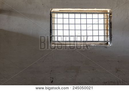 Old White Wall With Window Covered By Iron Grate. Sun Shining  Through Grill. Prison Room Interior .