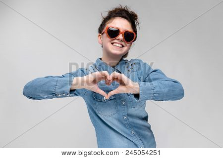 Young Girl With Glasses In Heart On Gray Background. Portrait Of A Young Beautiful Girl. Charming Gi